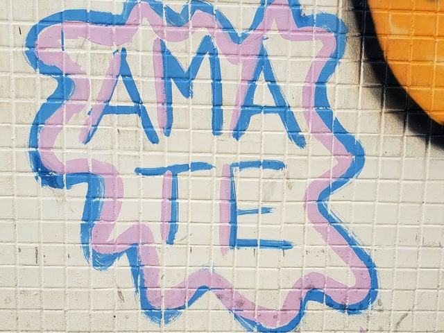 A palavra AMA-TE grafitada em azul e rosa sobre um muro de azulejos brancos.
