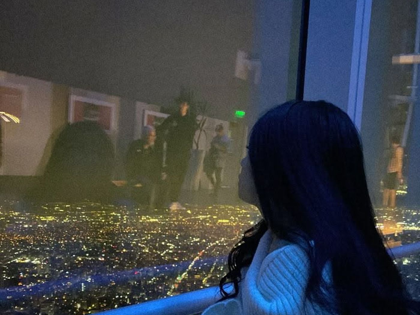 Mulher de costas, abraçada a si mesma, diante das luzes de uma cidade à noite.