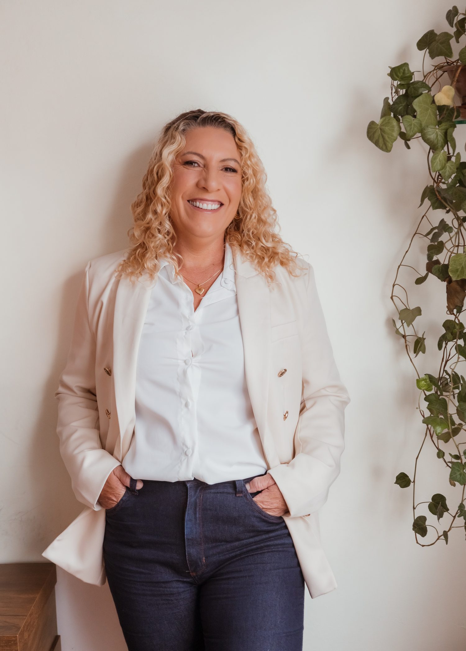 Viviana Prado, psicóloga clínica, em pé e sorrindo, de blazer claro e mãos nos bolsos, ao lado de uma planta.